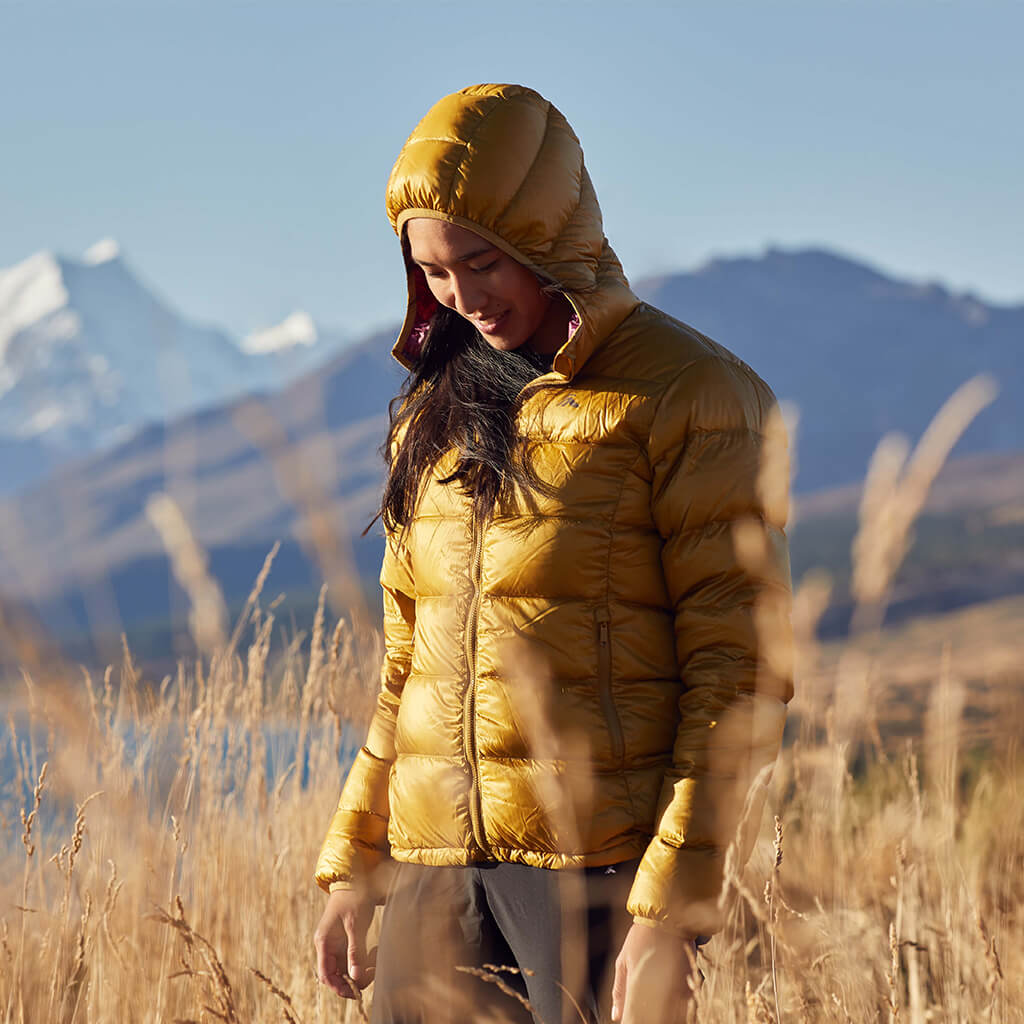 A woman wearing a mustard coloured macpac jupiter jacket with the hood up on sunny day