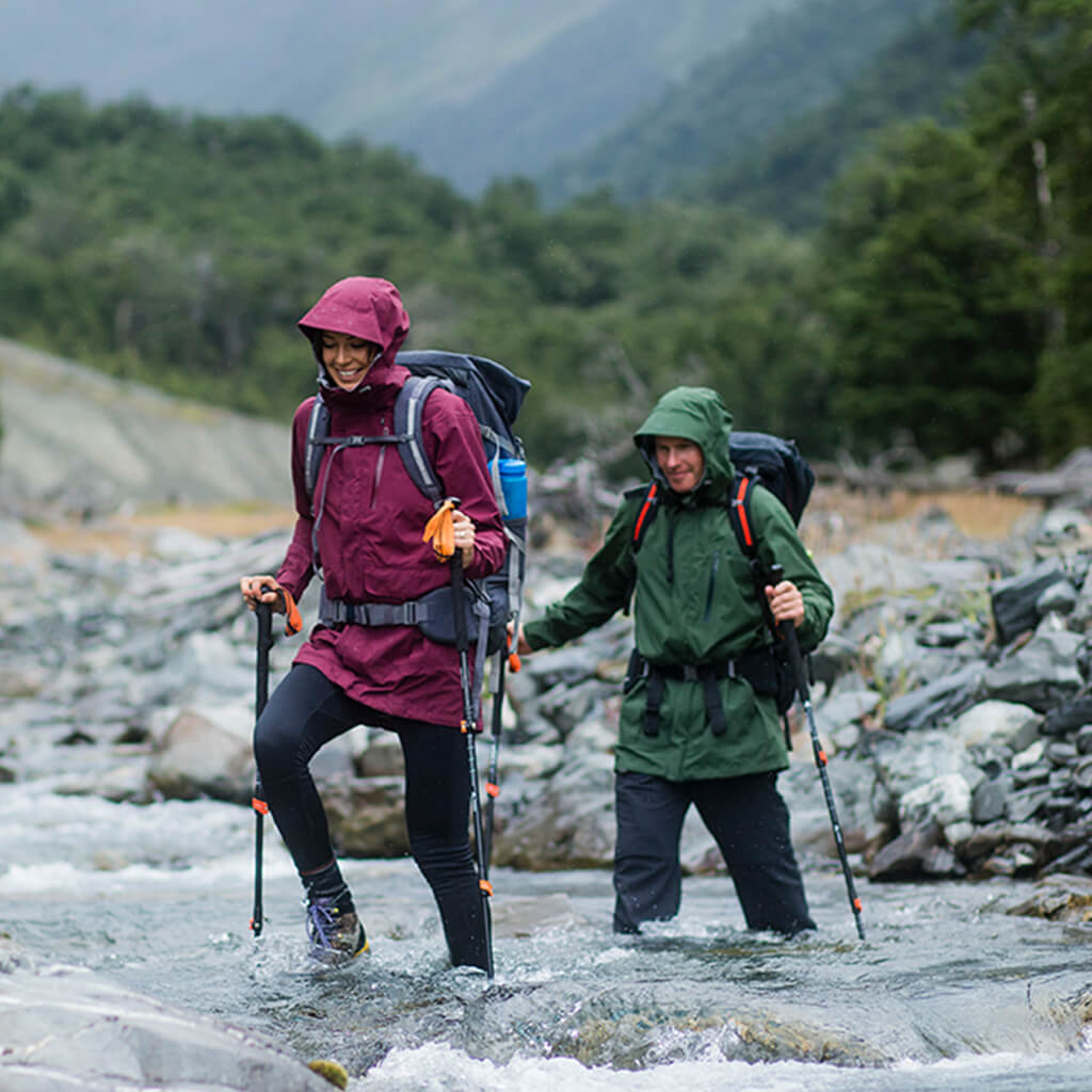 A man and a woman wearing hooded rain jackets hiking through a river using poles