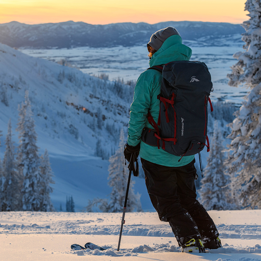 Skier standing on mountain at sunset wearing turquoise jacket and macpac backpack