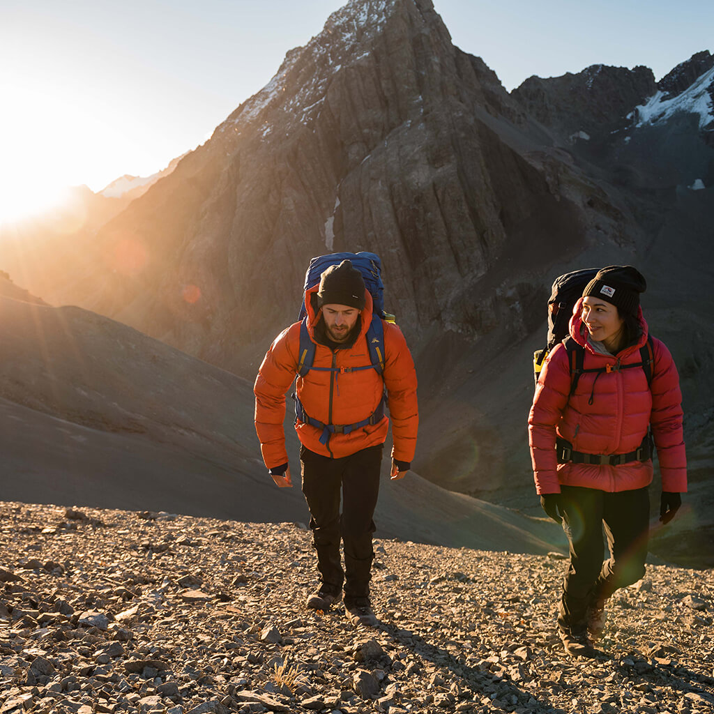 2 people wearing insulated jackets hiking a steep mountain with snow in the background