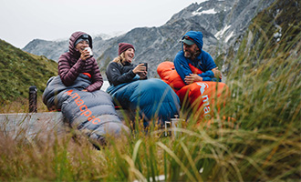 3 people sitting outside in sleeping bags with grass surrounding them