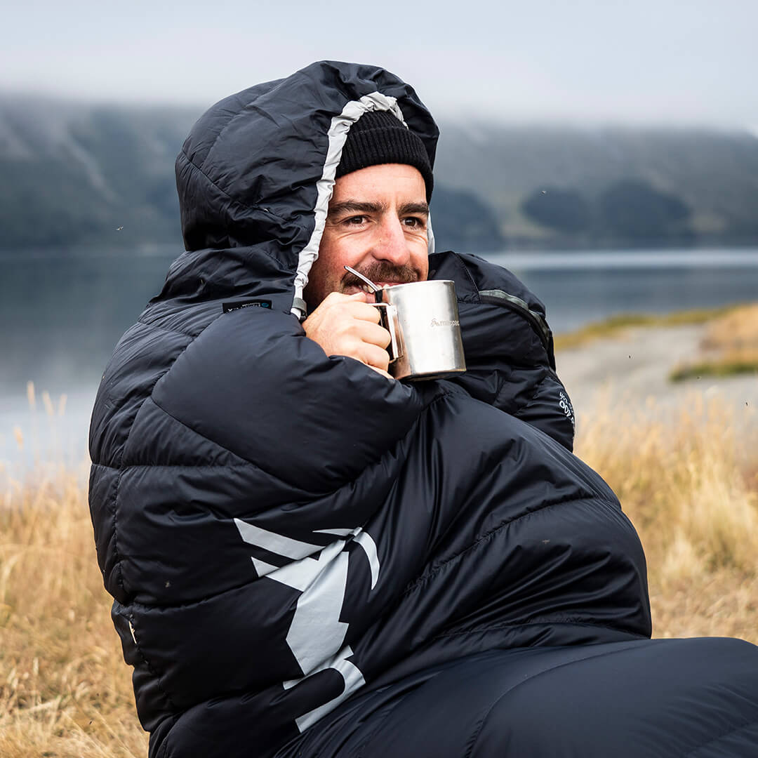Man sitting outside snuggled in a black macpac sleeping bag sipping a hot drink