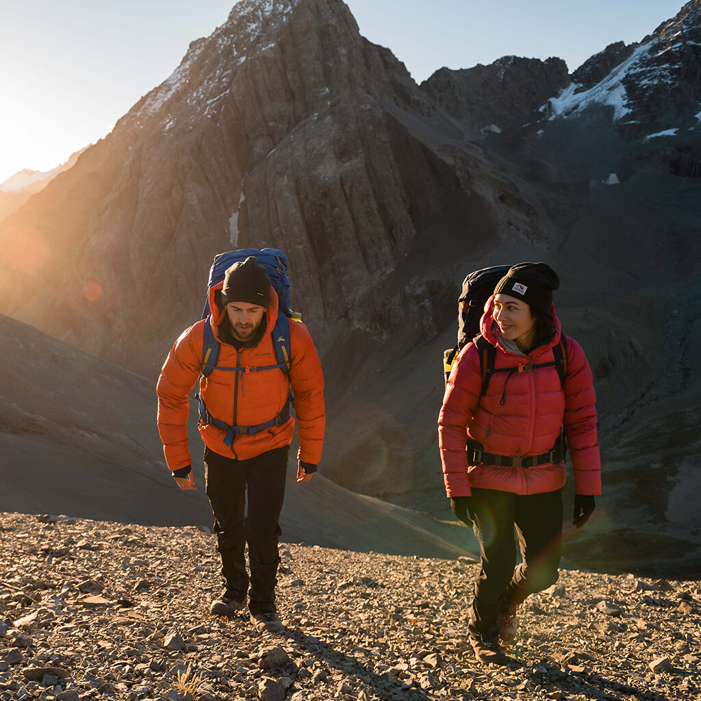 A man and a woman wearing hooded rain jackets hiking through a river using poles