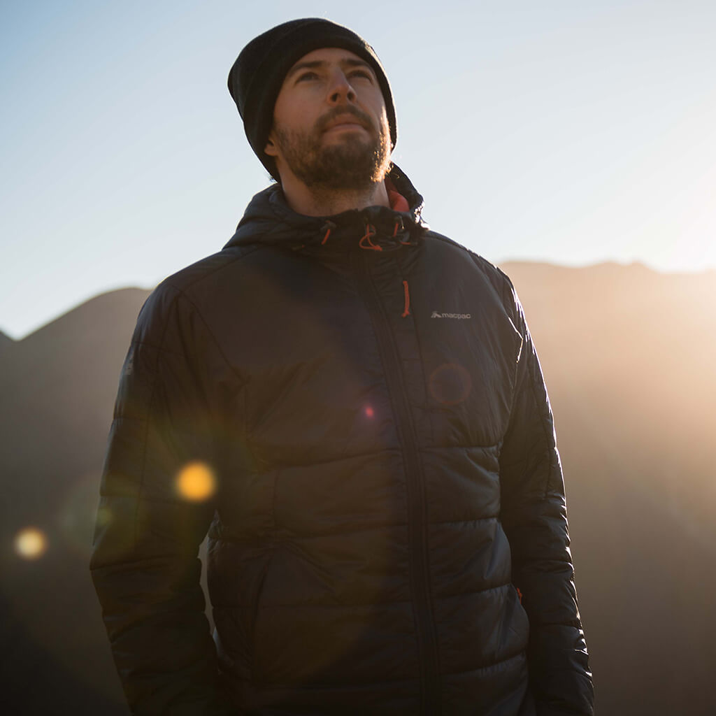 A man wearing a black macpac puffer jacket with a black beanie in front of mountain range on sunny day